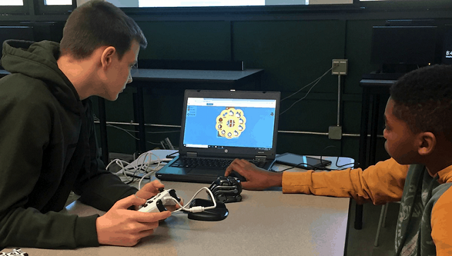 Two boys use a controller to navigate a program on a laptop sitting in front of them on the table