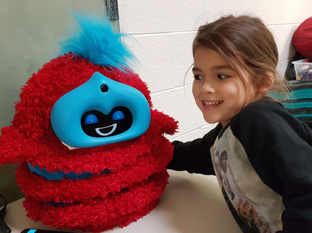 Girl smiles at a smiling fluffy red Tega robot with its arms outstretched.