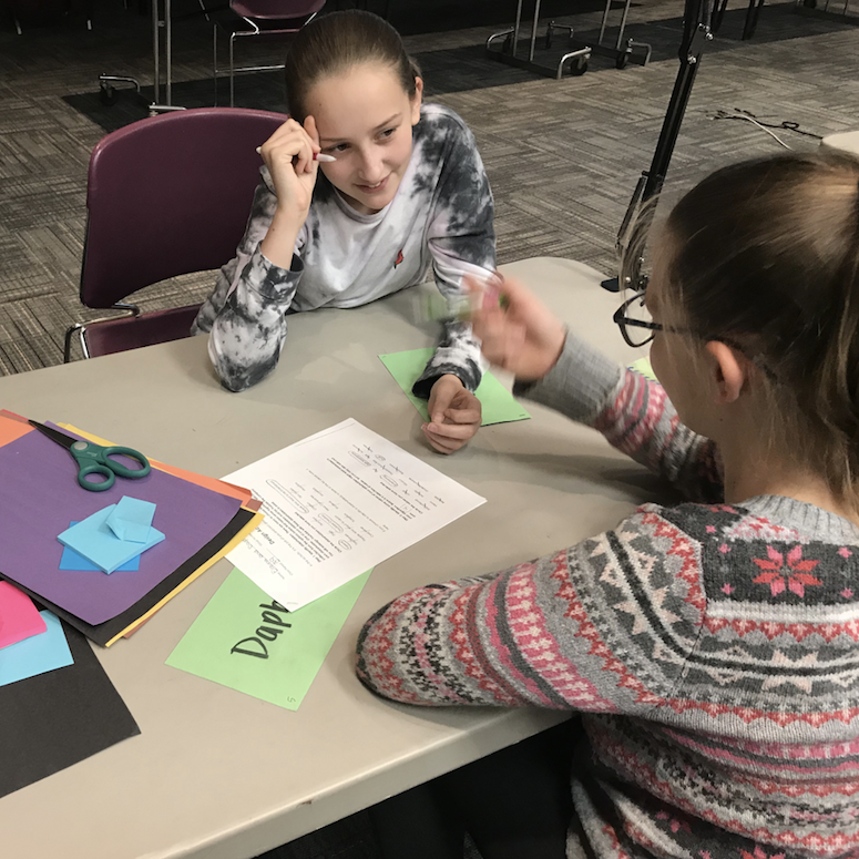 Two girls discuss a worksheet while sitting across from each other at a table.