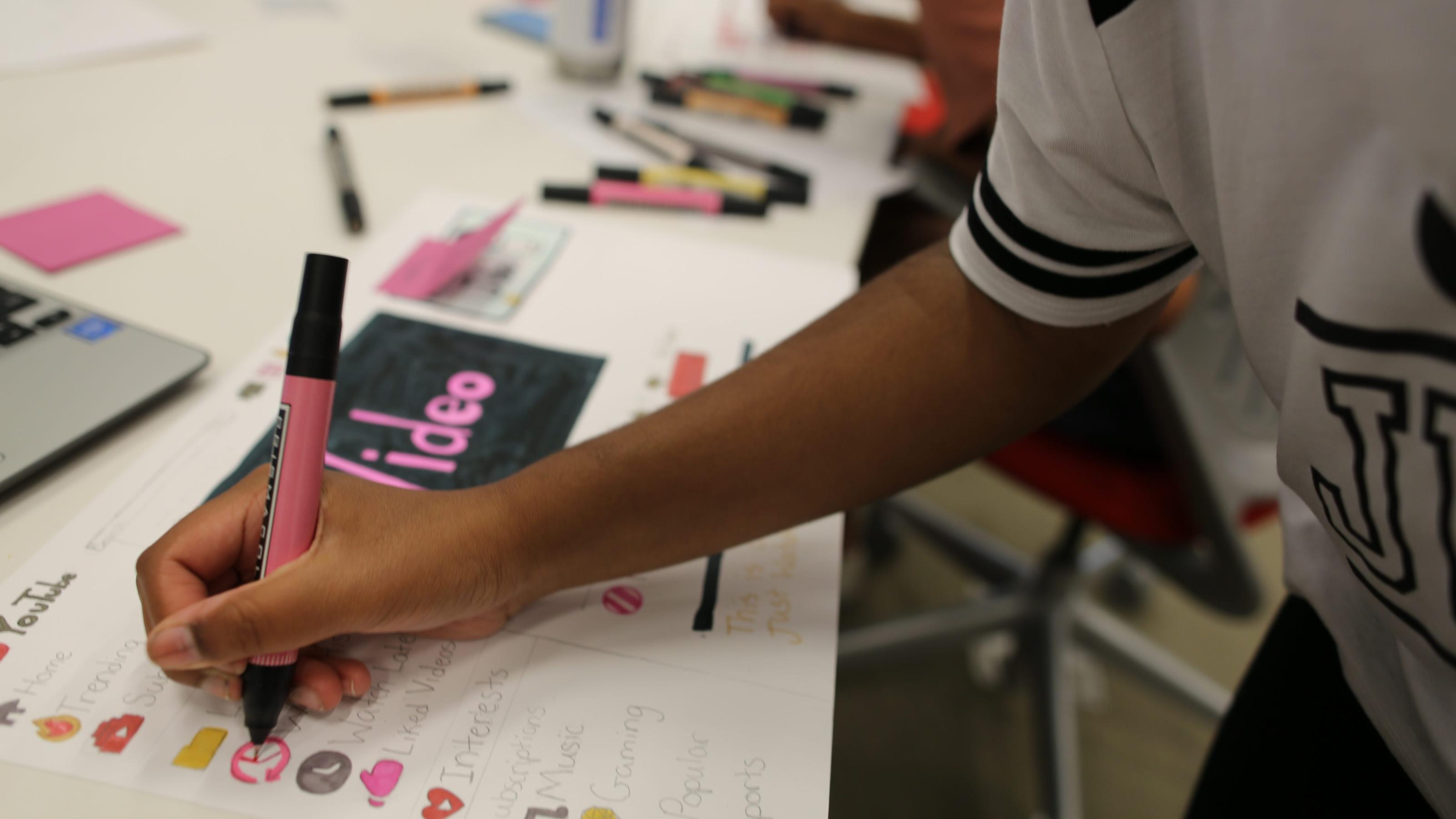 Child with a pink marker draws on a poster on which they have redesigned the YouTube user interface.